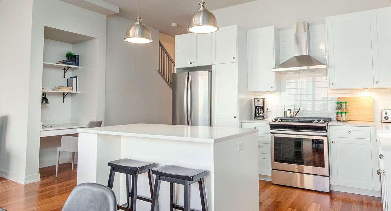 Modern apartment kitchen and discreet home office nook at The Ardus in Chicago, featuring white cabinets, an island, and pendant lighting