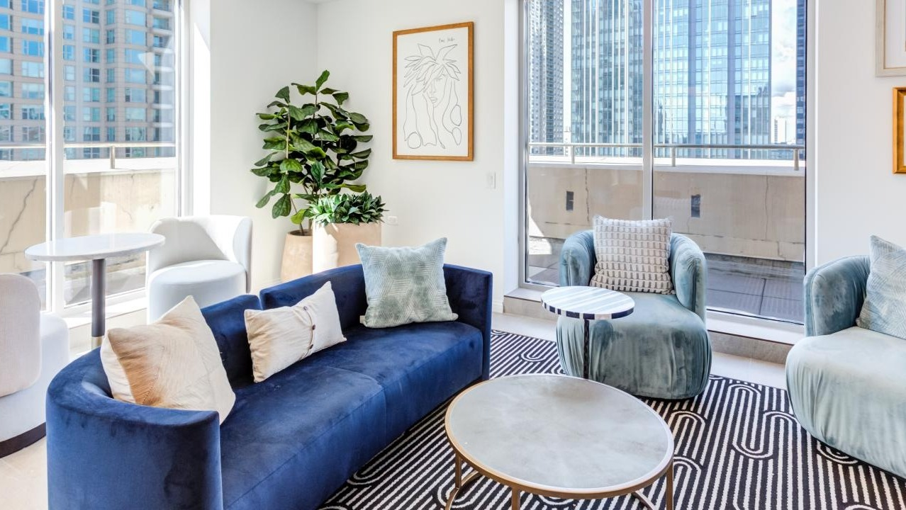 Vibrant resident lounge featuring a deep blue curved sofa, plush chairs, and large windows with city views at The Bernardin Apartments in Chicago