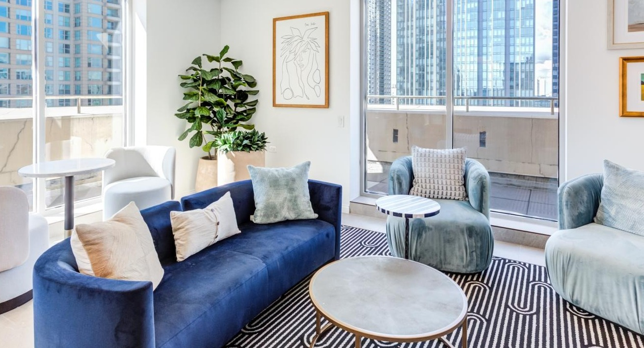 Vibrant resident lounge featuring a deep blue curved sofa, plush chairs, and large windows with city views at The Bernardin Apartments in Chicago