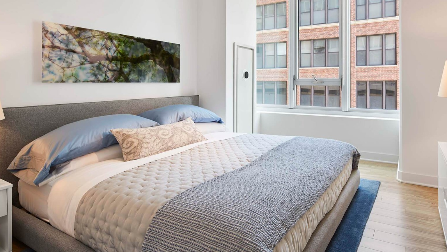 Cozy bedroom with a queen-sized bed, soft blue linens, stylish lamp, and a large window overlooking the city at The Cooper in Chicago