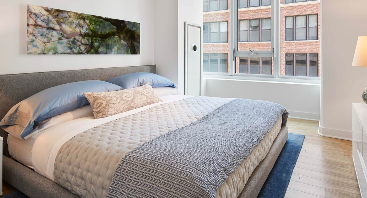 Cozy bedroom with a queen-sized bed, soft blue linens, stylish lamp, and a large window overlooking the city at The Cooper in Chicago