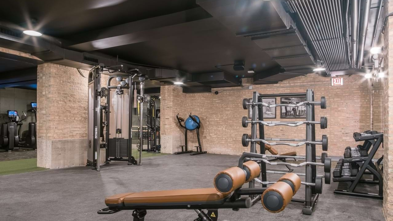 Comprehensive weight training gym with racks of free weights, benches, and various machines, all within an exposed brick setting at The Duncan in Chicago