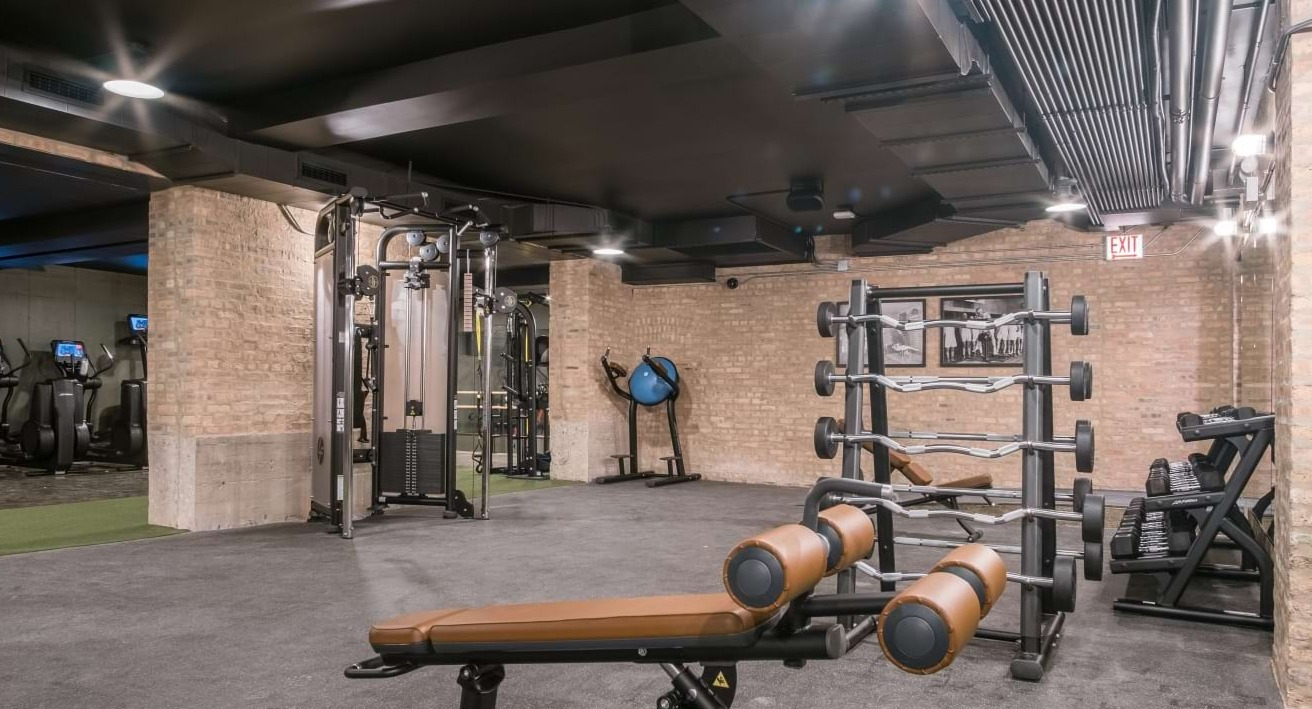 Comprehensive weight training gym with racks of free weights, benches, and various machines, all within an exposed brick setting at The Duncan in Chicago