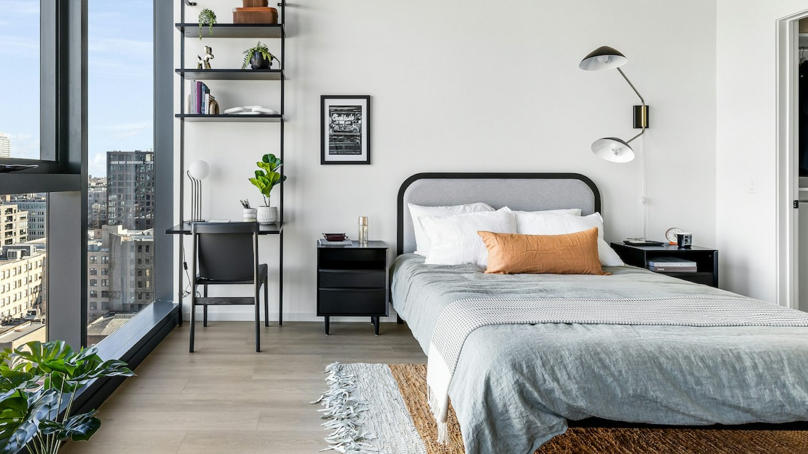 Contemporary bedroom featuring a comfortable bed, functional workspace with shelving, and large windows offering expansive city views at The Dylan in Chicago