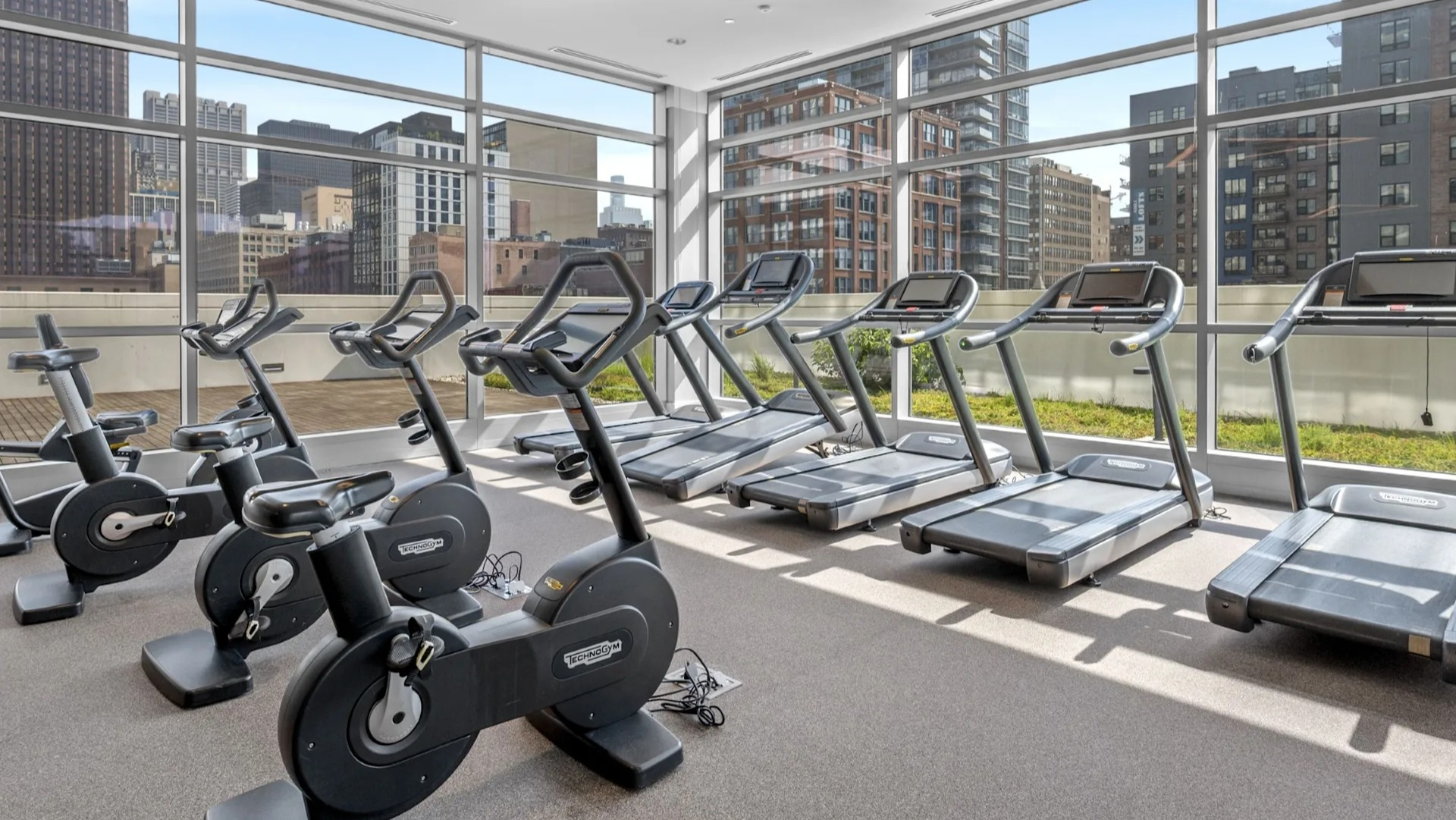 Bright cardio fitness room with a selection of treadmills and stationary bikes, offering city views through large windows at The Elle in Chicago