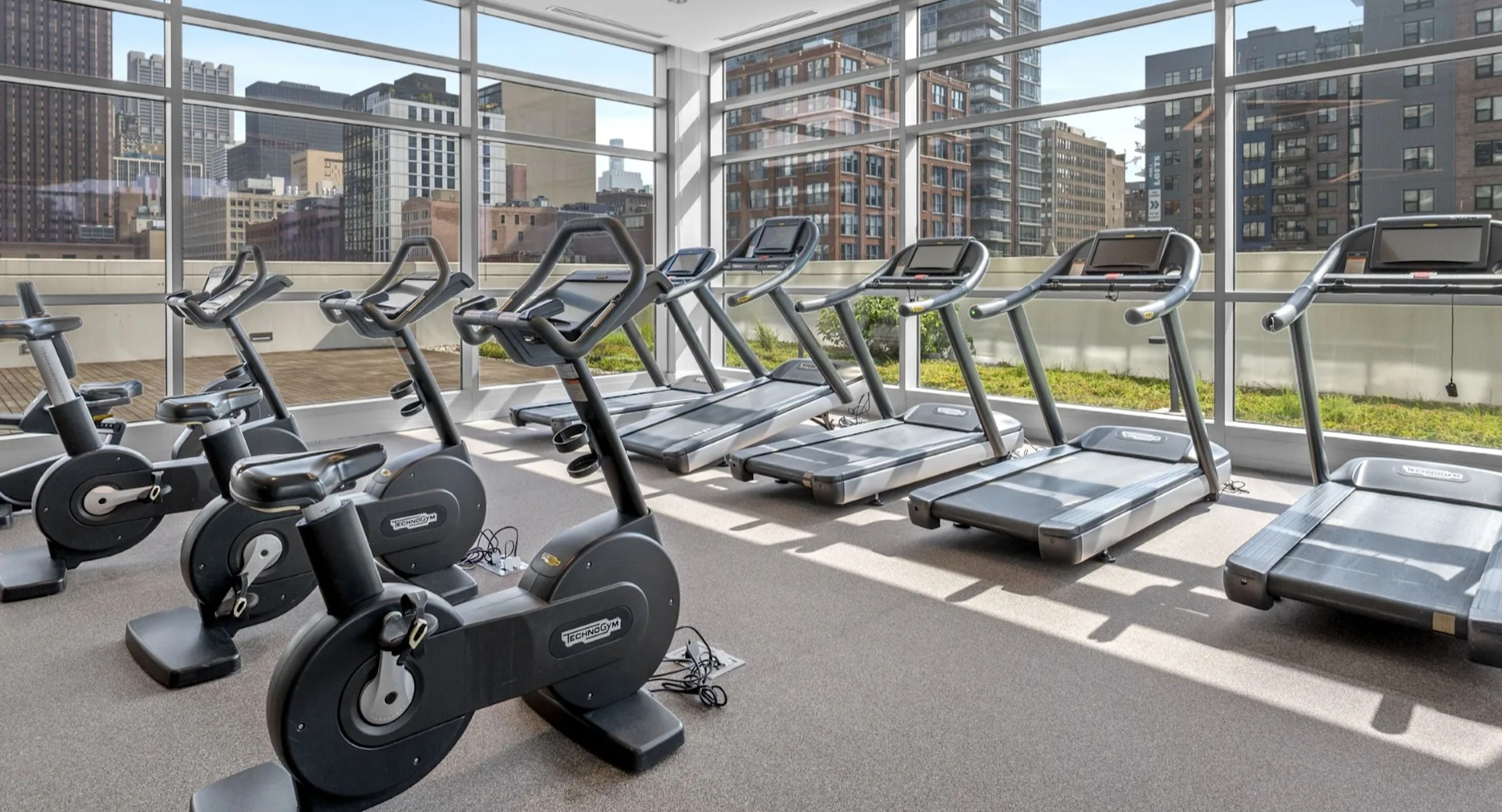 Bright cardio fitness room with a selection of treadmills and stationary bikes, offering city views through large windows at The Elle in Chicago