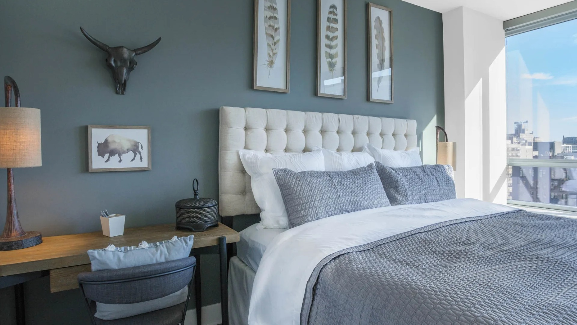 Designer bedroom with a tufted headboard, accent wall, dedicated workspace, and large windows offering city views at The Elle in Chicago