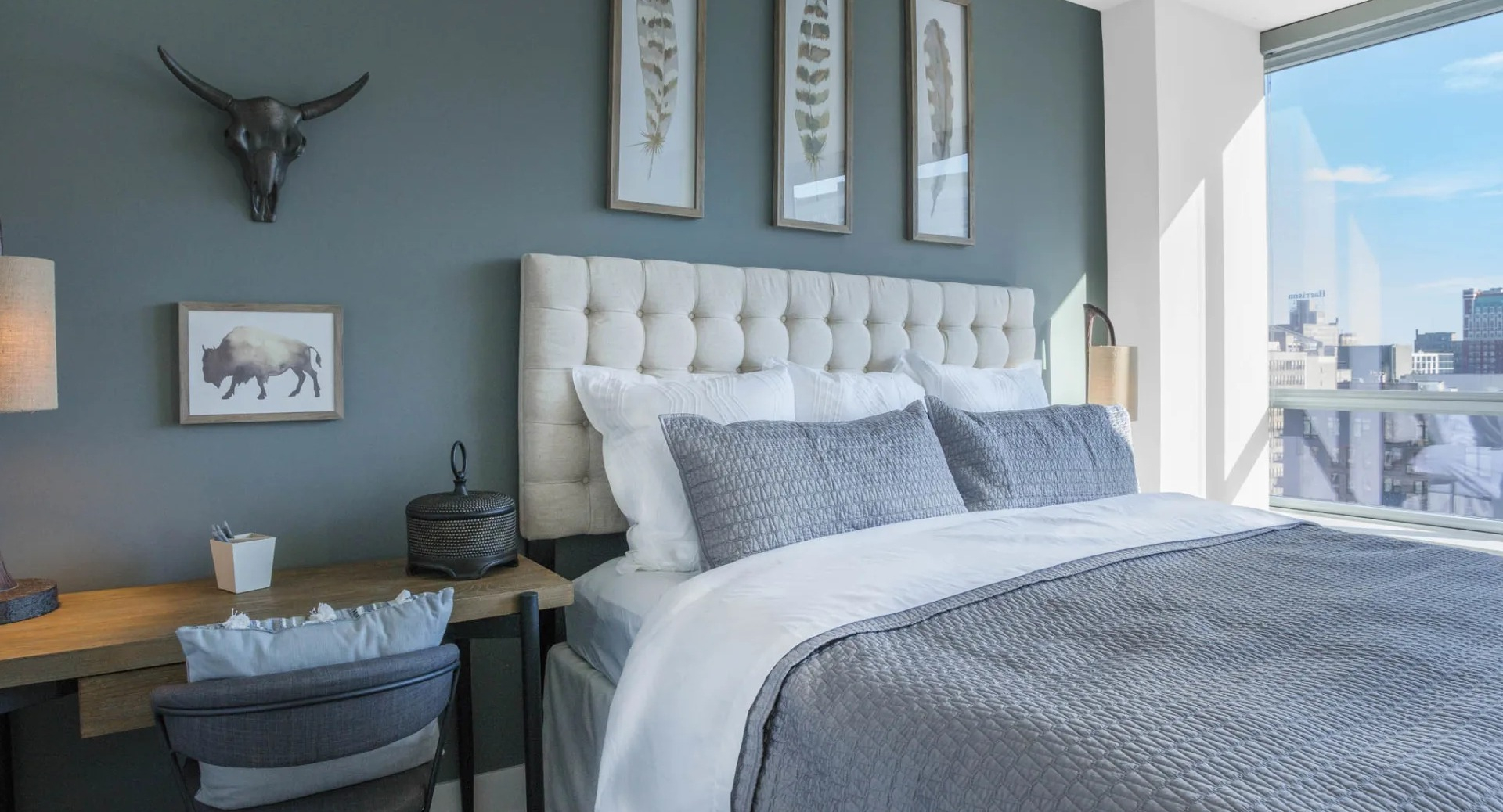 Designer bedroom with a tufted headboard, accent wall, dedicated workspace, and large windows offering city views at The Elle in Chicago