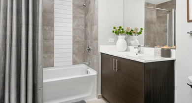 A modern apartment bathroom featuring a white bathtub with a gray-tiled shower, a vanity, and a large mirror at The Madison at Racine in Chicago