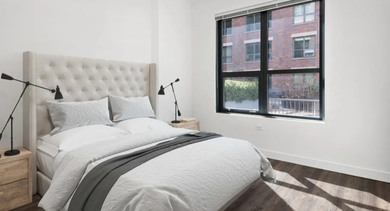 A cozy bedroom with a tufted headboard, crisp white bedding, wooden nightstands, and a large window at The Madison at Racine in Chicago