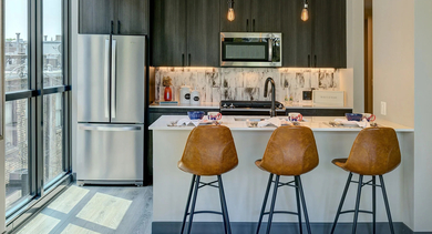 A sleek, modern kitchen with dark cabinetry, stainless steel appliances, a white island with bar stools, and a large window overlooking a balcony at The Mason