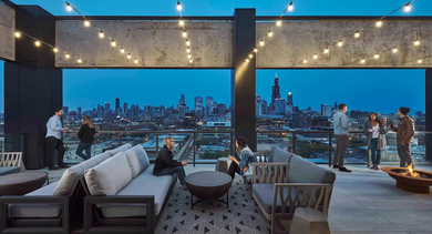 People socializing on a vibrant rooftop deck at dusk, featuring lounge seating, a fire pit, string lights, and a stunning Chicago skyline view at The Mason