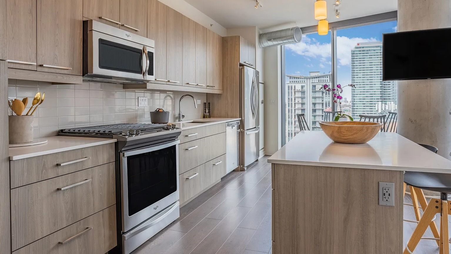 A bright and modern apartment kitchen with light wood cabinetry, stainless steel appliances, a kitchen island with seating, and ample natural light at The Parker Fulton Market