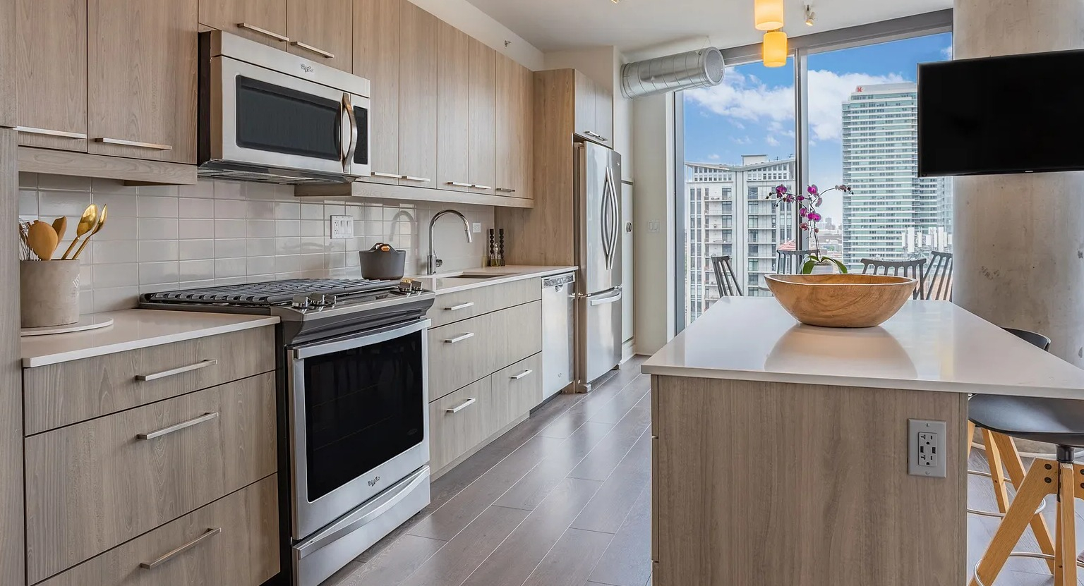 A bright and modern apartment kitchen with light wood cabinetry, stainless steel appliances, a kitchen island with seating, and ample natural light at The Parker Fulton Market