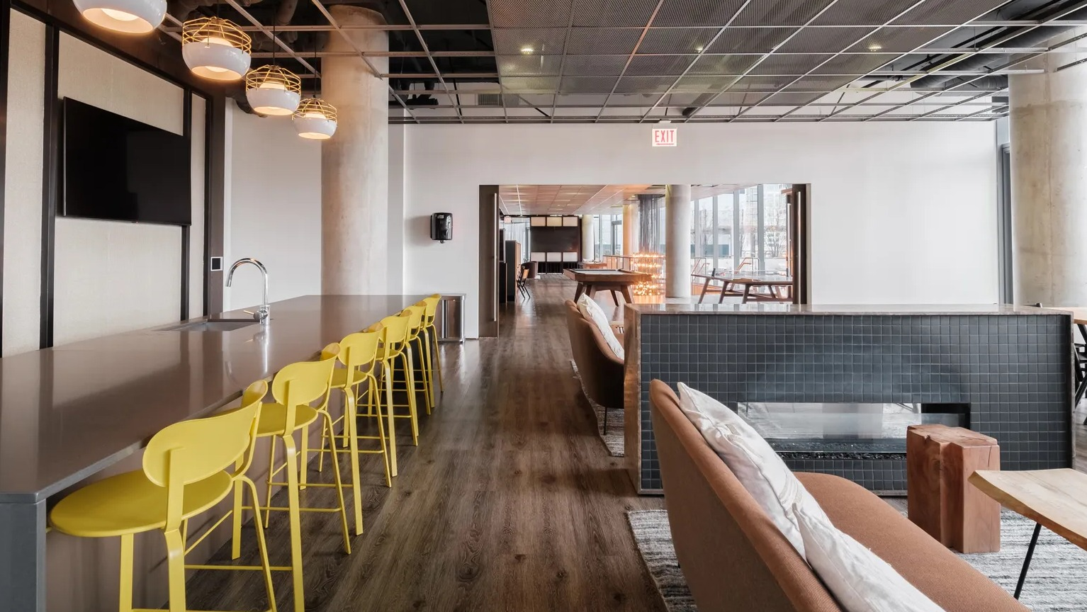 A stylish clubroom with a long bar, modern yellow stools, a fireplace, and comfortable lounge seating for residents at The Parker Fulton Market in Chicago