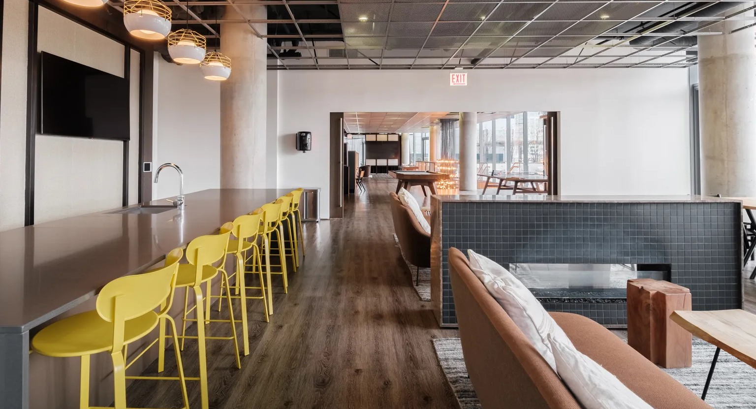 A stylish clubroom with a long bar, modern yellow stools, a fireplace, and comfortable lounge seating for residents at The Parker Fulton Market in Chicago