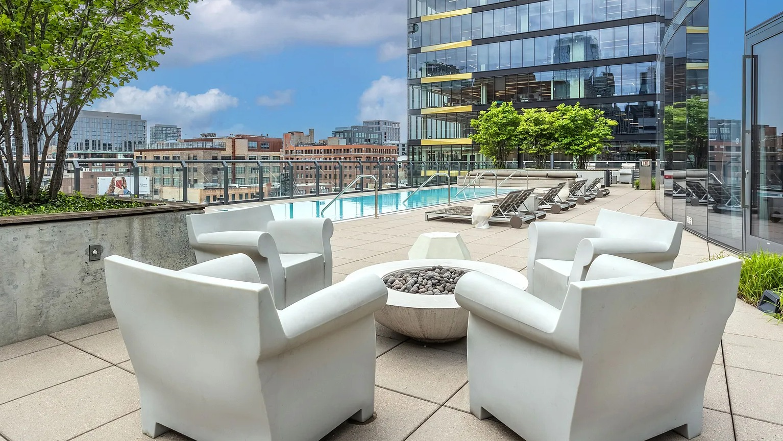 A chic pool deck with comfortable white lounge chairs, a modern fire pit, lush greenery, and the building's glass facade at The Parker Fulton Market in Chicago