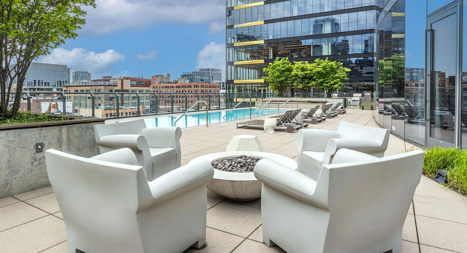 A chic pool deck with comfortable white lounge chairs, a modern fire pit, lush greenery, and the building's glass facade at The Parker Fulton Market in Chicago