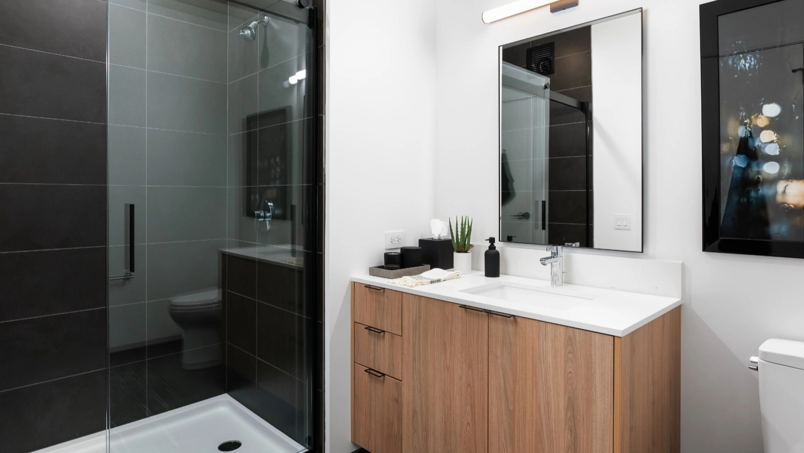 A sleek, modern bathroom featuring a spacious walk-in shower with dark tiling, a light wood vanity, and contemporary fixtures at The Reed in Chicago