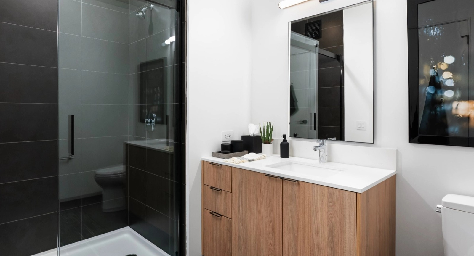 A sleek, modern bathroom featuring a spacious walk-in shower with dark tiling, a light wood vanity, and contemporary fixtures at The Reed in Chicago