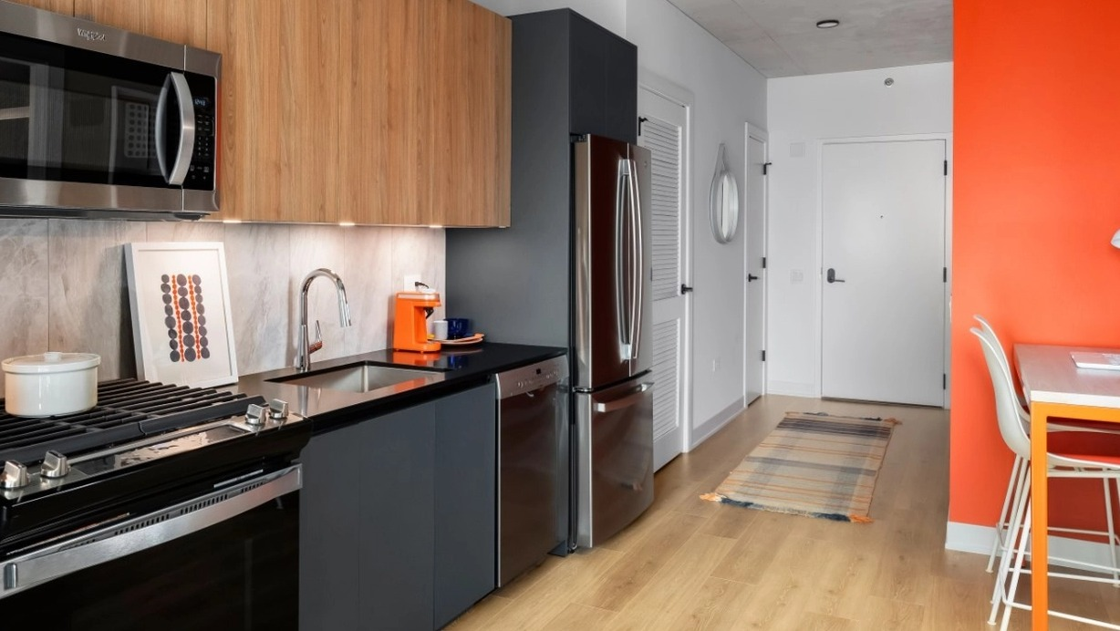 A contemporary kitchen with wooden cabinets, stainless steel appliances, and a vibrant orange accent wall with a functional desk area at The Reed in Chicago