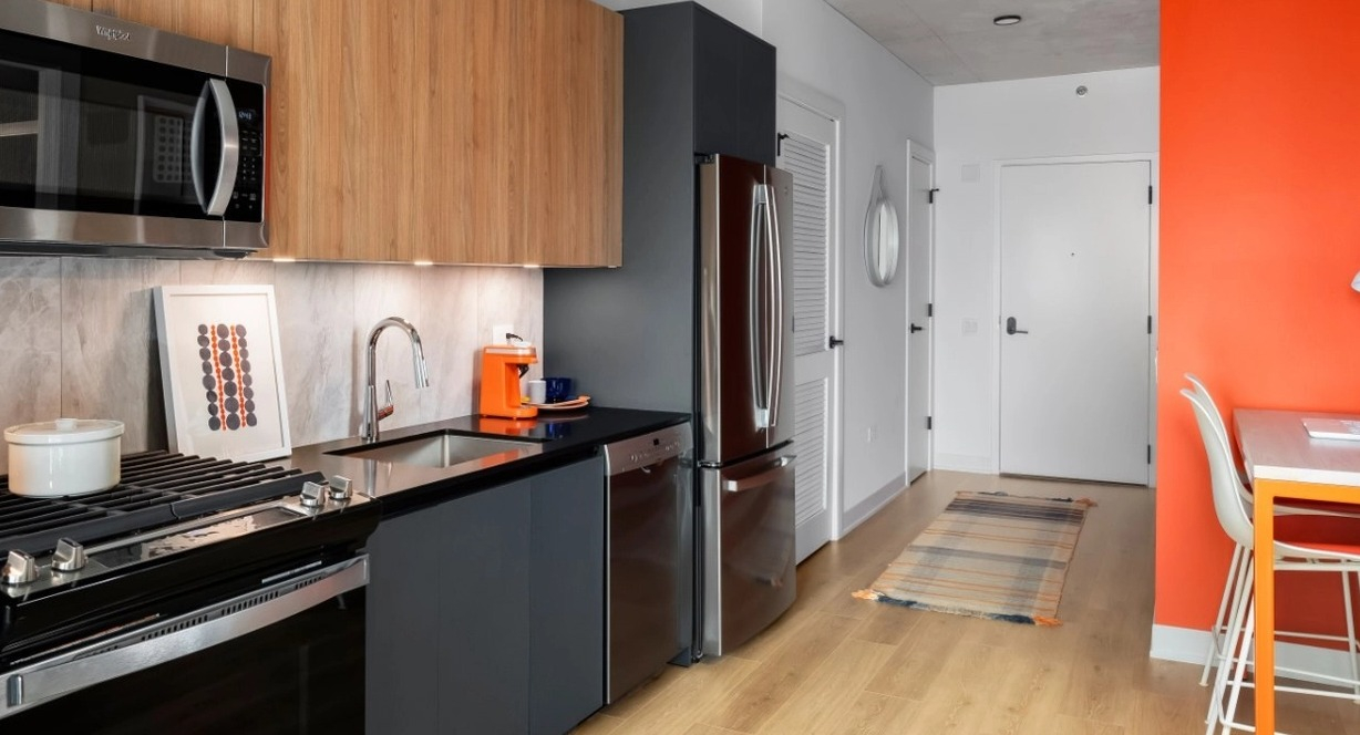 A contemporary kitchen with wooden cabinets, stainless steel appliances, and a vibrant orange accent wall with a functional desk area at The Reed in Chicago