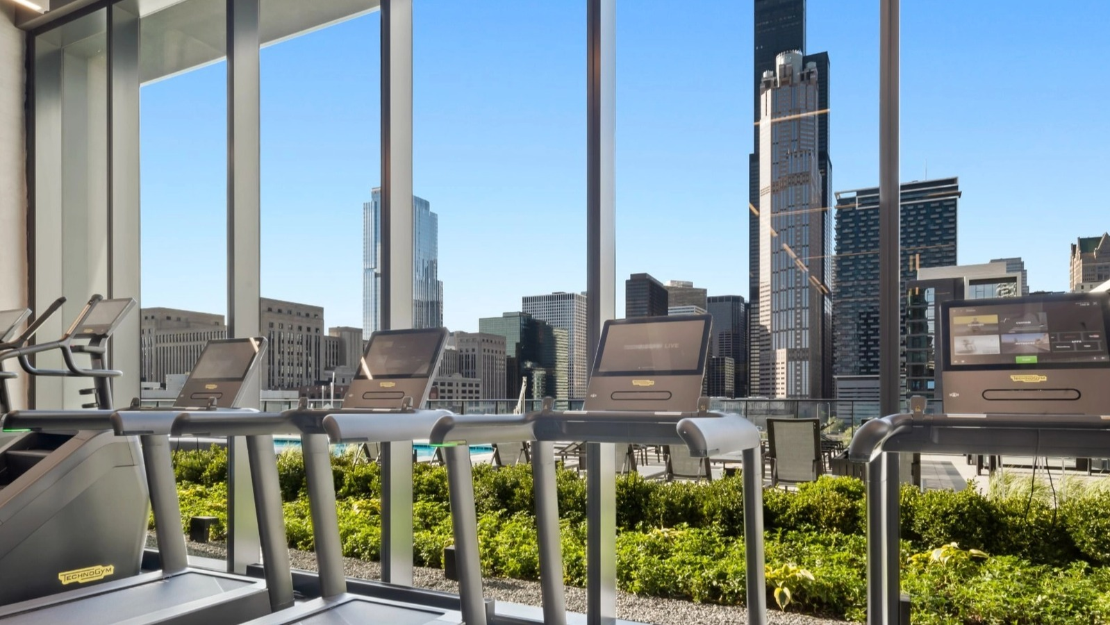 A state-of-the-art fitness center with treadmills and cardio equipment, featuring large windows with panoramic Chicago skyline views at The Reed