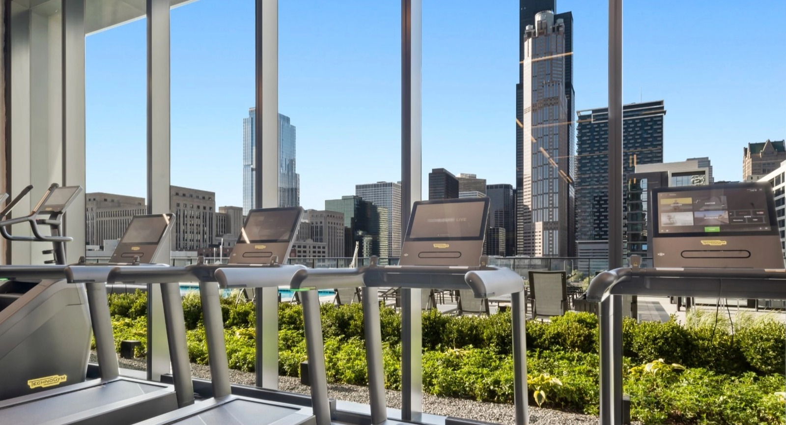 A state-of-the-art fitness center with treadmills and cardio equipment, featuring large windows with panoramic Chicago skyline views at The Reed