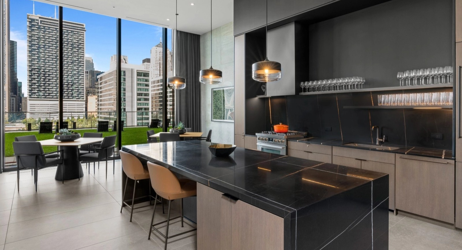 A luxurious modern kitchen with dark cabinetry, a large island, and a dining area, all with expansive city views from floor-to-ceiling windows at The Reed in Chicago