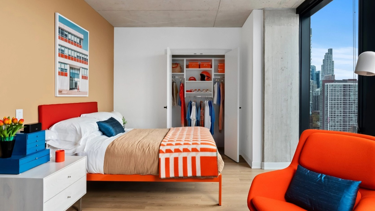 A modern and vibrant bedroom with colorful bedding, an orange accent chair, an open closet, and a large window showcasing urban views at The Reed in Chicago