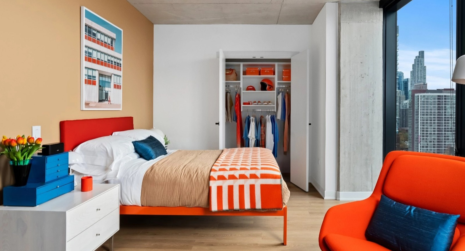 A modern and vibrant bedroom with colorful bedding, an orange accent chair, an open closet, and a large window showcasing urban views at The Reed in Chicago