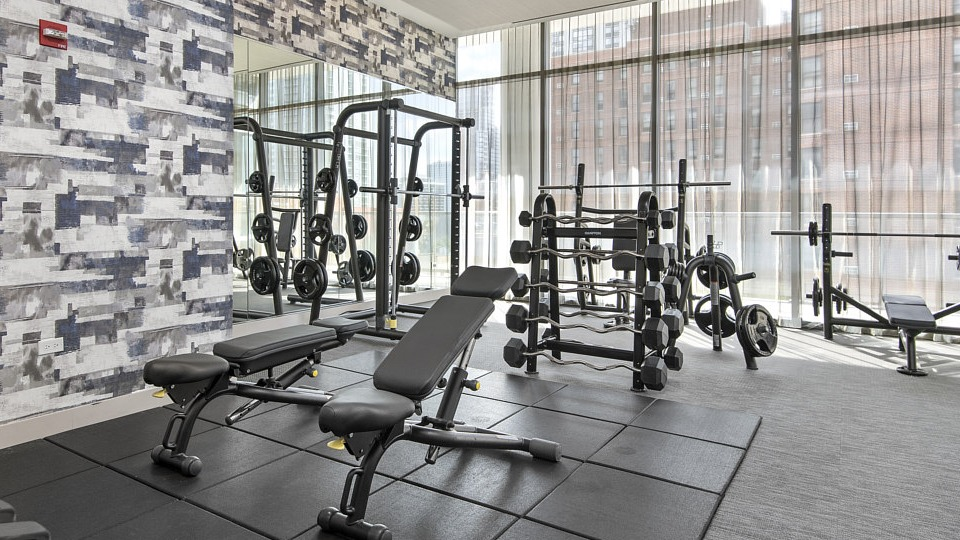 A modern fitness center with ample space for strength training, featuring free weights, benches, and power racks at The Sinclair in Chicago