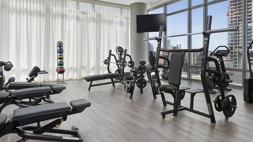 A bright fitness center with various strength training equipment, weights, and cardio machines, all with expansive city views at The Sinclair in Chicago