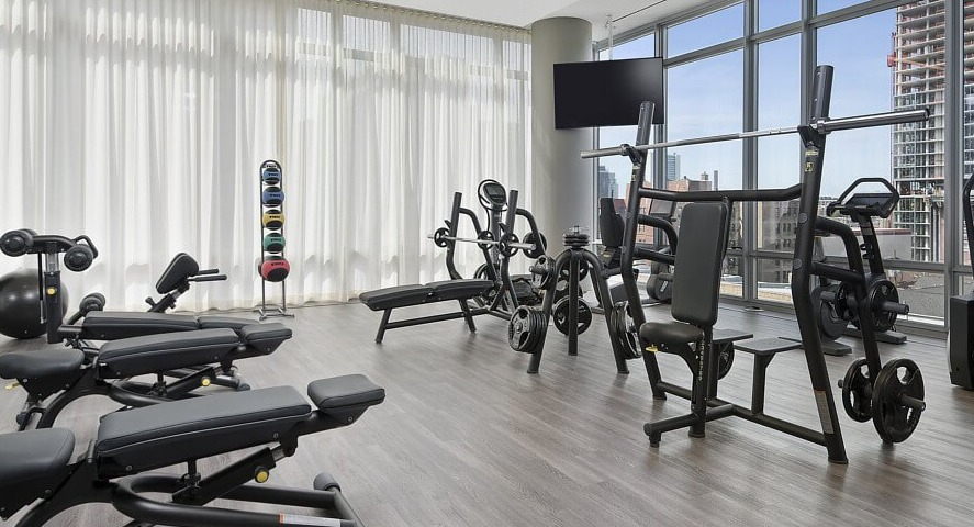 A bright fitness center with various strength training equipment, weights, and cardio machines, all with expansive city views at The Sinclair in Chicago