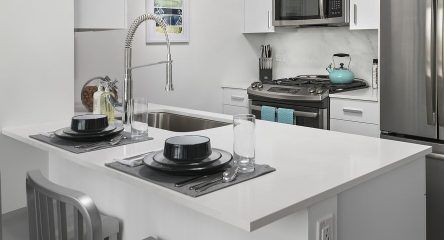 A close-up of a modern kitchen island with bar seating, white countertops, and stainless steel appliances at The Sinclair apartments in Chicago