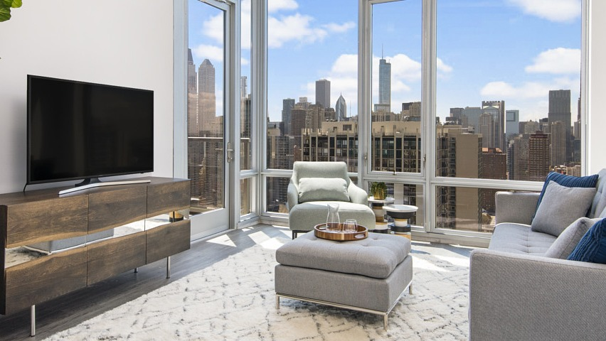 A bright apartment living room with a large sofa, TV console, and floor-to-ceiling windows offering expansive Chicago city views at The Sinclair