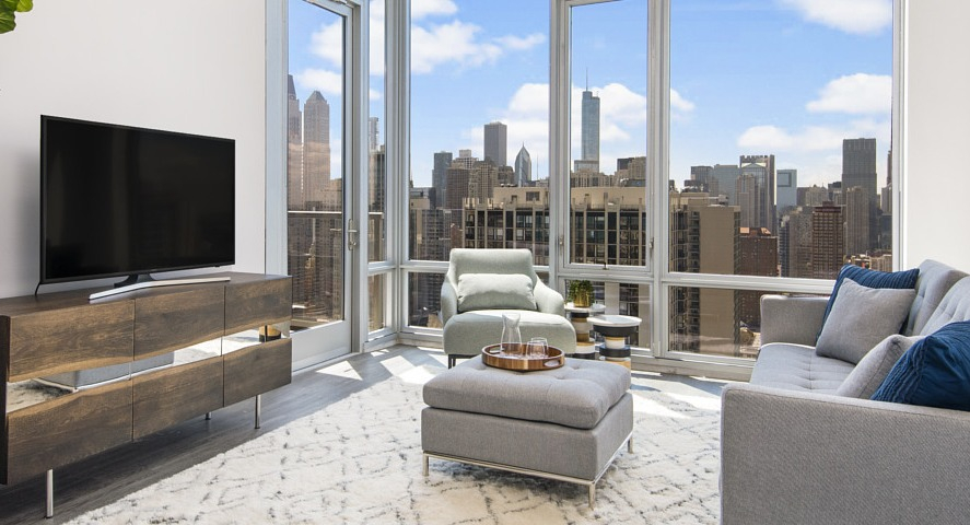 A bright apartment living room with a large sofa, TV console, and floor-to-ceiling windows offering expansive Chicago city views at The Sinclair