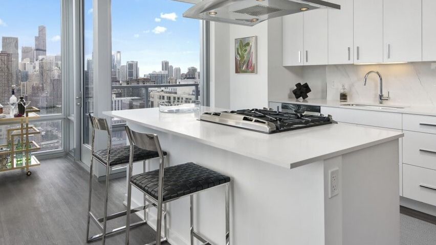A modern white kitchen with a large island, stainless steel appliances, and stunning city views from floor-to-ceiling windows at The Sinclair in Chicago