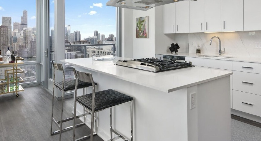 A modern white kitchen with a large island, stainless steel appliances, and stunning city views from floor-to-ceiling windows at The Sinclair in Chicago