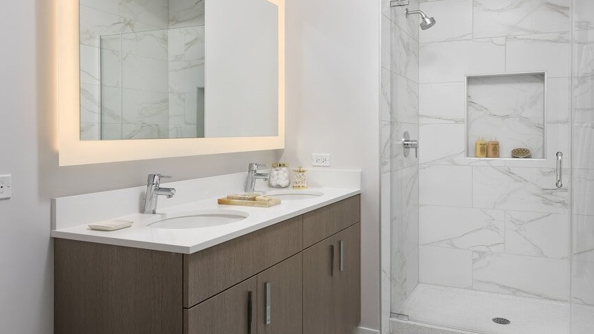 A luxurious master bathroom with a double vanity, a large lighted mirror, and a spacious walk-in shower with elegant tile at The Sinclair in Chicago