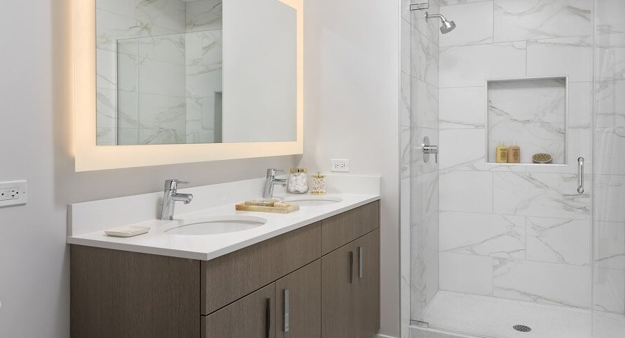 A luxurious master bathroom with a double vanity, a large lighted mirror, and a spacious walk-in shower with elegant tile at The Sinclair in Chicago