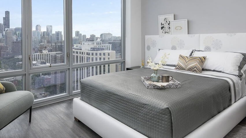 A chic and comfortable bedroom with a modern bed, elegant headboard, and expansive city skyline views through large windows at The Sinclair in Chicago