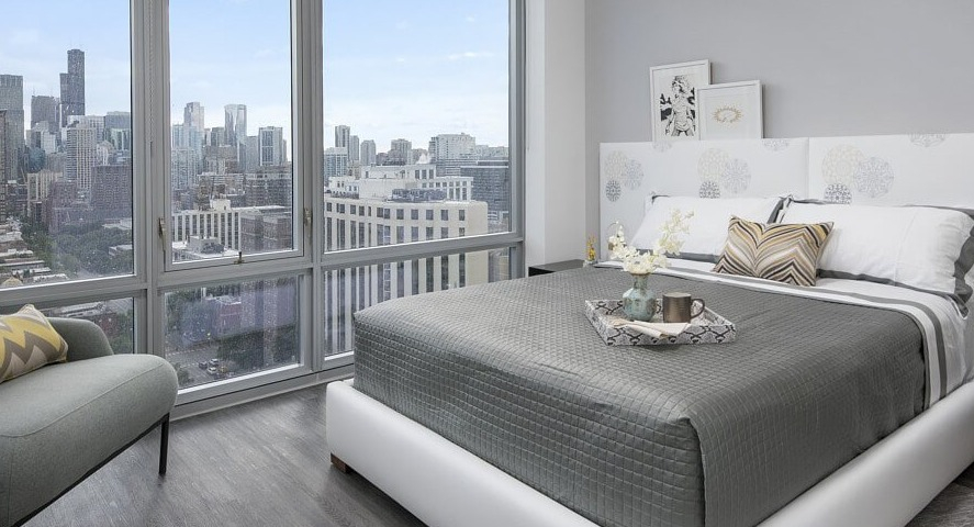 A chic and comfortable bedroom with a modern bed, elegant headboard, and expansive city skyline views through large windows at The Sinclair in Chicago
