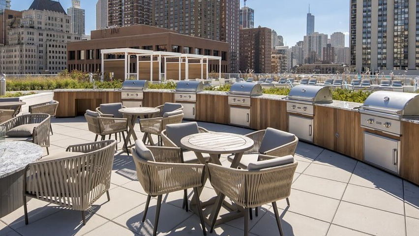 An inviting rooftop patio with multiple grilling stations, round dining tables, and a modern fire pit, offering city views at The Sinclair in Chicago