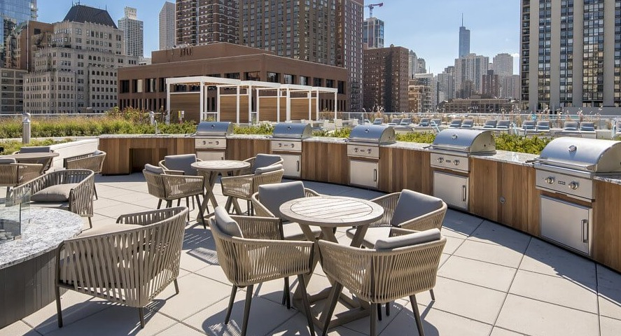 An inviting rooftop patio with multiple grilling stations, round dining tables, and a modern fire pit, offering city views at The Sinclair in Chicago