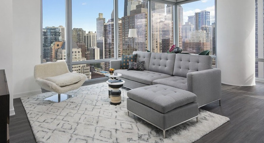 A luxurious living room with a large sectional sofa, elegant rug, and floor-to-ceiling windows offering panoramic city skyline views at The Sinclair in Chicago