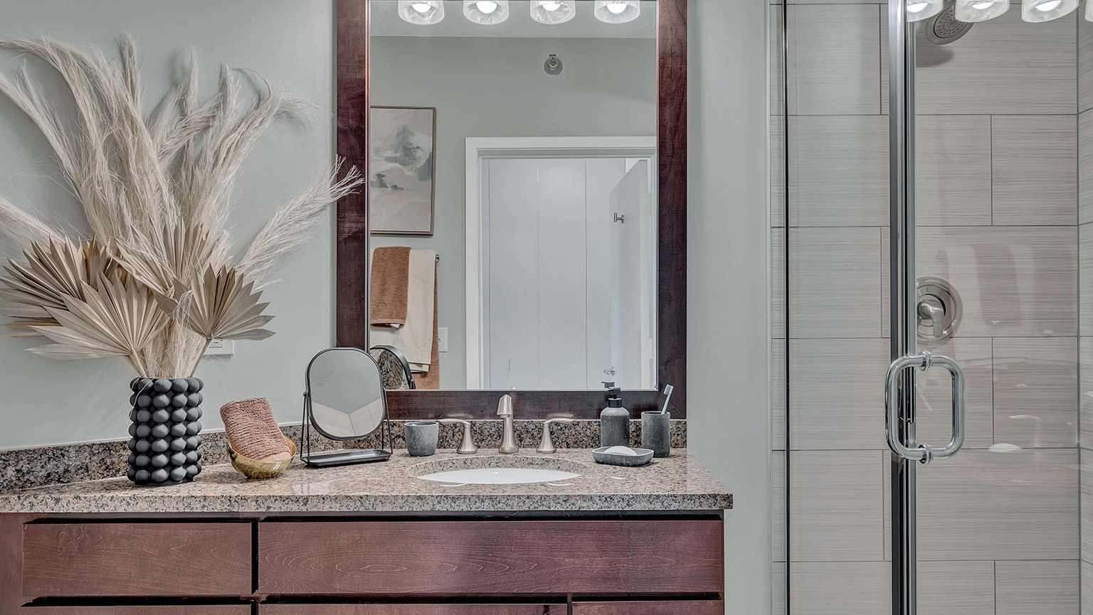 A modern bathroom featuring a spacious vanity with granite countertops, a large mirror, and a sleek walk-in shower at The Streeter in Chicago