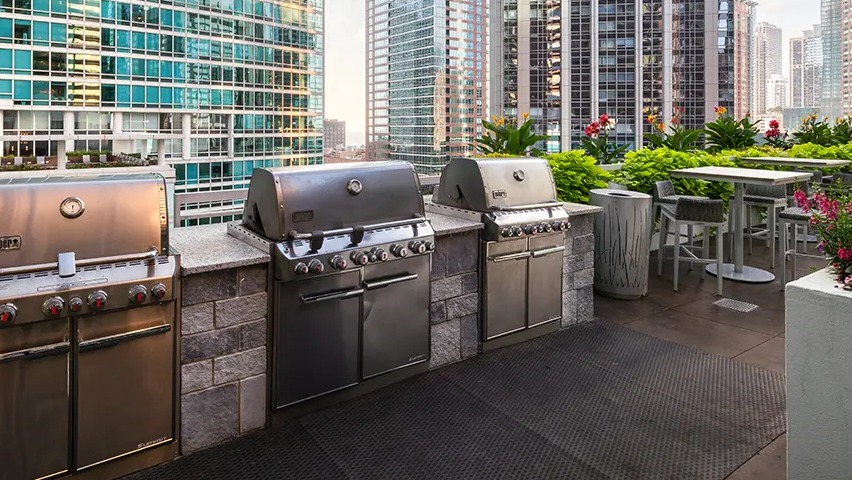 A modern outdoor grilling area with multiple high-end grills and counter space, set against a stunning cityscape backdrop at The Streeter in Chicago