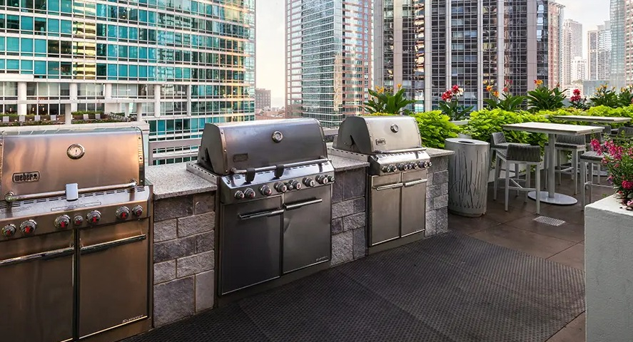 A modern outdoor grilling area with multiple high-end grills and counter space, set against a stunning cityscape backdrop at The Streeter in Chicago