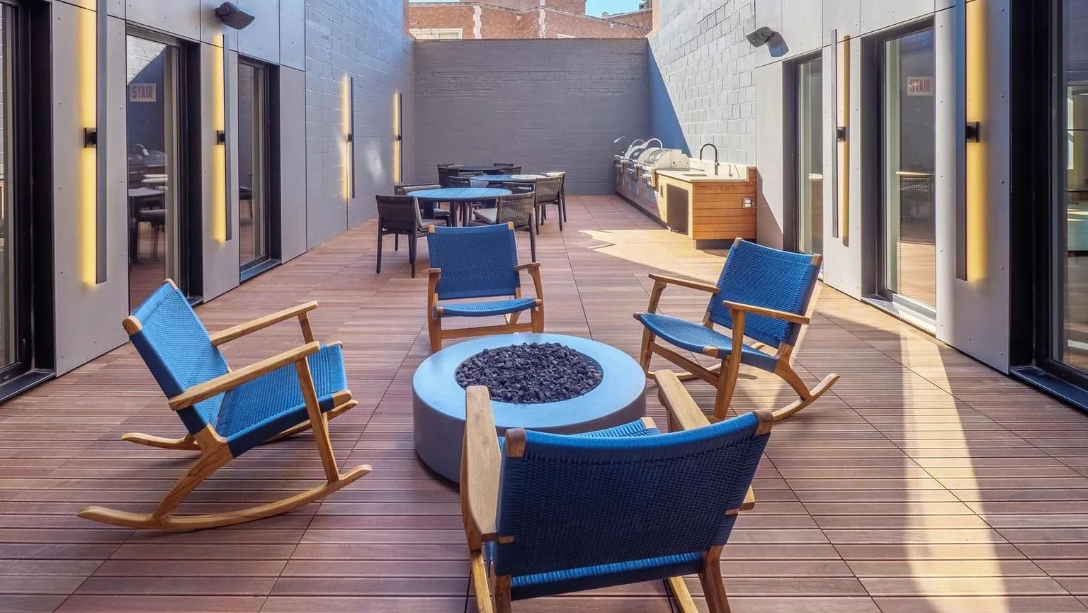 A chic outdoor patio featuring comfortable rocking chairs, a modern fire pit, and grilling stations, surrounded by brick buildings at The Thompson at Fulton Market in Chicago